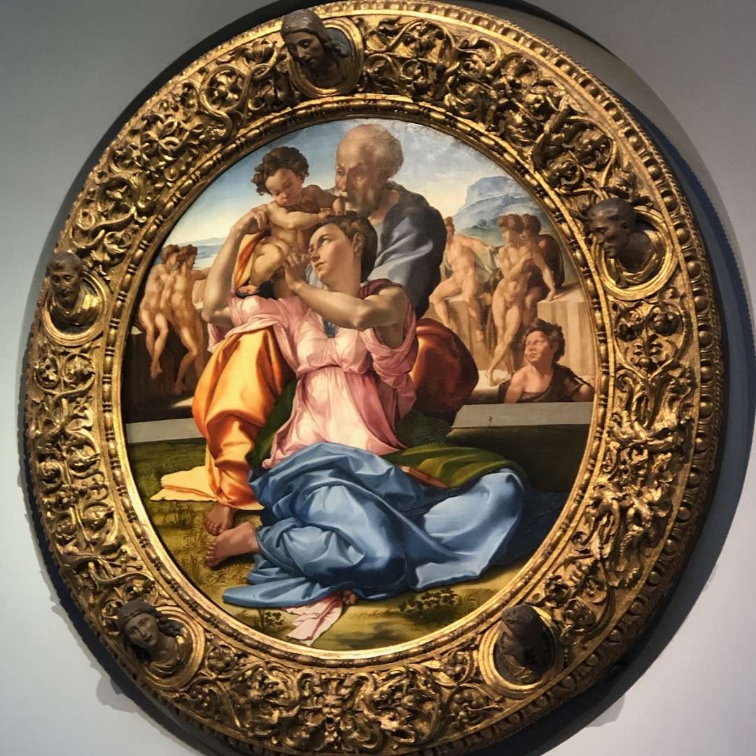 Round painting with elaborate frame of three figures in foreground and many naked people in the background at the UIffizi in Florence